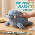 A handmade crocheted manatee doll in light blue-gray is shown on a wooden table with a human hand resting on its back. The image includes text overlays reading 'MY CUDDLY MANATEE FRIEND' and 'HANDMADE AMIGURUMI CROCHET DOLL'.