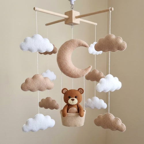 A felt baby mobile with a wooden ceiling mount, featuring a large tan crescent moon, white and tan clouds, and a brown teddy bear sitting in a small basket.