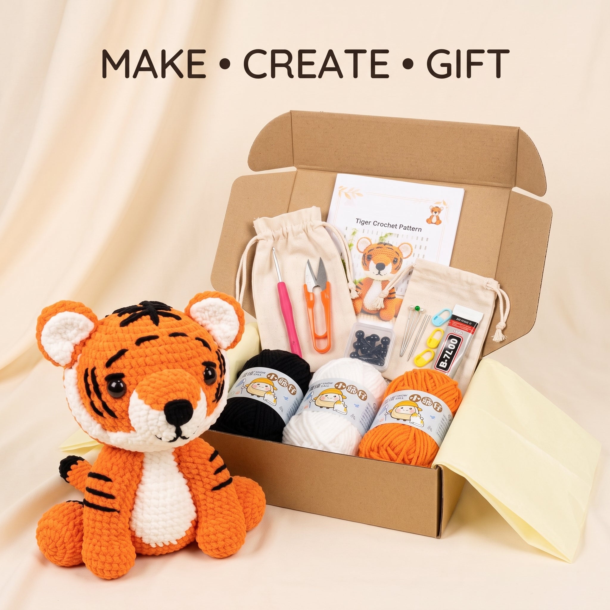 A tiger-themed crochet kit displayed in an open cardboard box with a crocheted tiger toy beside it. The kit includes yarn balls, a drawstring bag, a pattern card, tools, and accessories, with the text 'MAKE • CREATE • GIFT' above on a warm beige background.