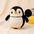 A crocheted penguin toy with a black and white body, orange beak and feet, standing on a soft cream fabric background with yarn balls blurred in the back.