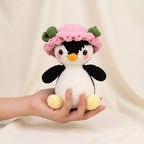 A crocheted penguin doll with a pink flower hat is held in a person's hand against a soft beige fabric backdrop.