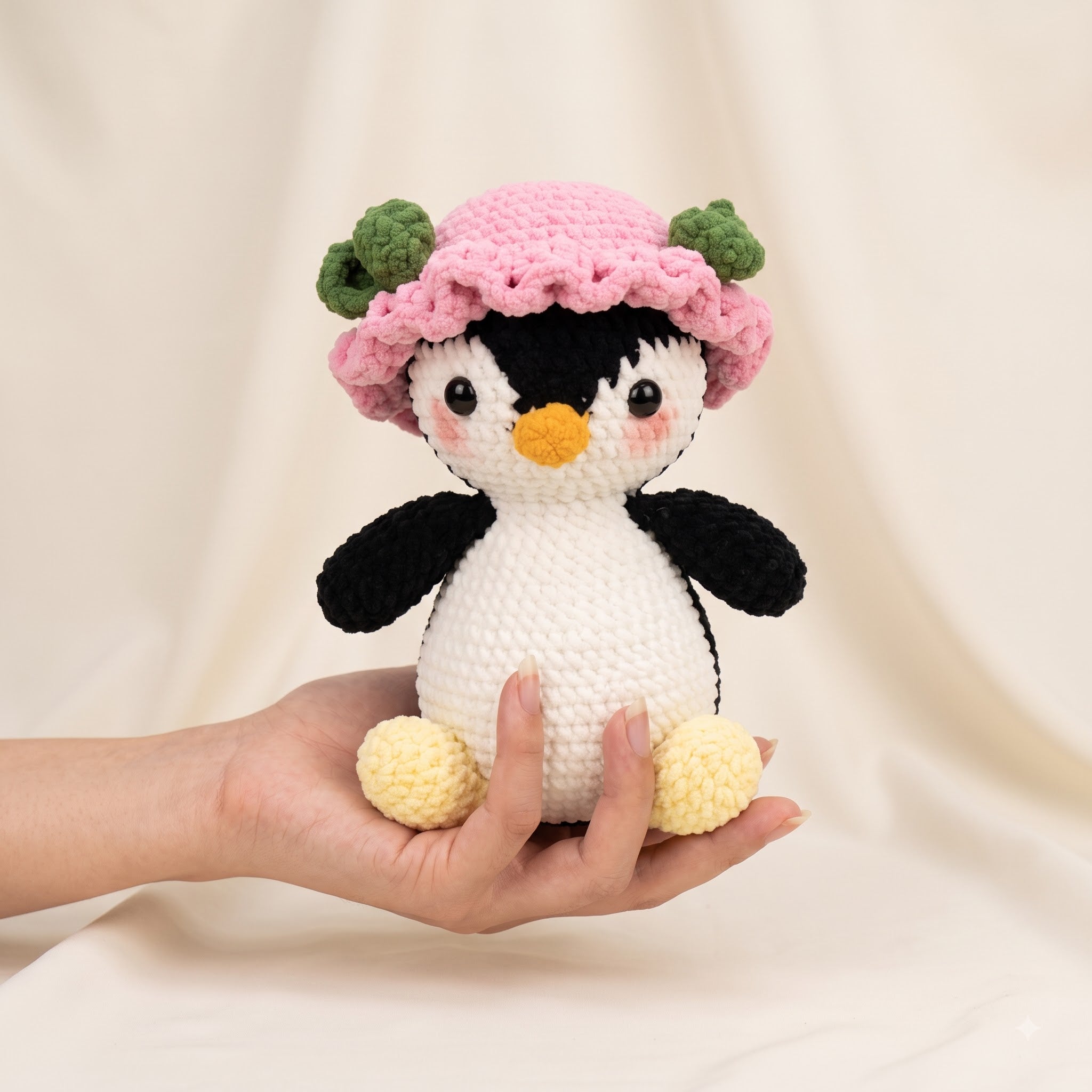 A crocheted penguin doll with a pink flower hat is held in a person's hand against a soft beige fabric backdrop.