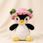 A crocheted penguin toy wearing a pink hat with green accents, posed against a soft beige fabric backdrop.