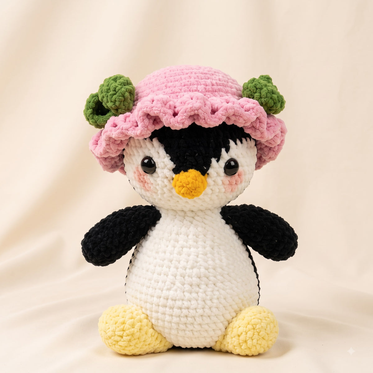 A crocheted penguin toy wearing a pink hat with green accents, posed against a soft beige fabric backdrop.
