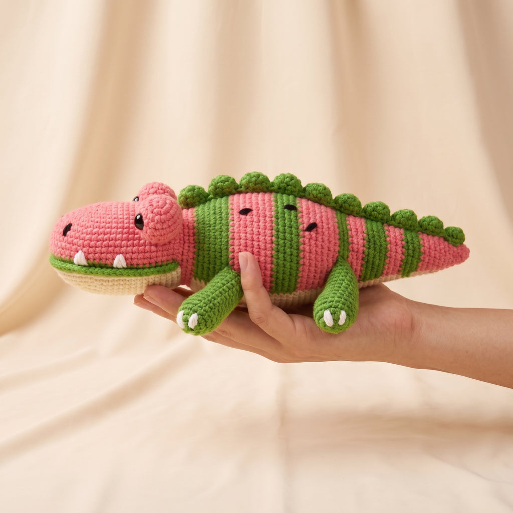 A crocheted plush crocodile toy with pink and green stripes, held in a person's hand against a soft beige fabric backdrop.