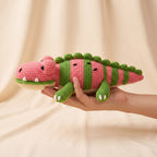 A crocheted plush crocodile toy with pink and green stripes, held in a person's hand against a soft beige fabric backdrop.