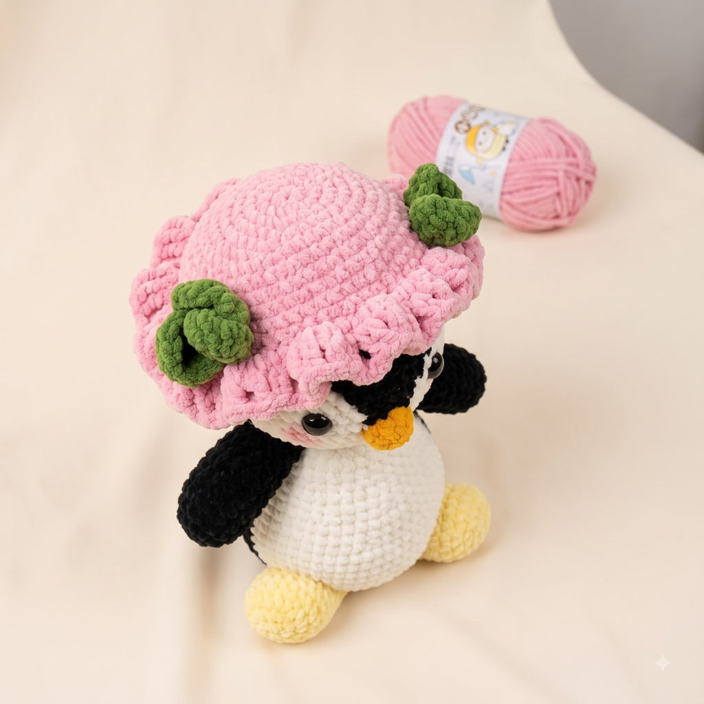 A crocheted penguin toy wearing a pink flower hat with green leaves, posed on a light beige fabric surface with a pink yarn ball in the background.