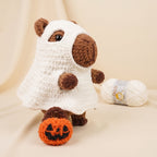 A crocheted capybara doll dressed as a ghost, holding an orange pumpkin, with a ball of white yarn in the background on a beige fabric backdrop.