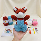 A crocheted fox doll wearing light blue overalls is held in a person's hand, with yarn balls, a pattern booklet, and small crafting tools arranged on a cream fabric backdrop.