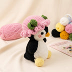 A crocheted penguin toy wearing a pink hat with green leaves, posed on a cream fabric backdrop with yarn balls and a card in the background.