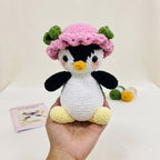A crocheted penguin doll being held in a person's hand, wearing a pink hat with green leaf accents, a yellow beak, black and white body, and yellow feet. A small booklet and yarn balls sit on the table in the background.
