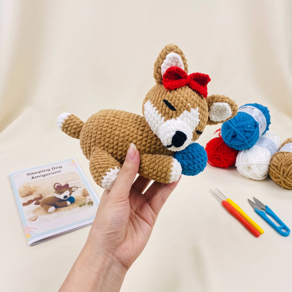 DIY Sleeping Dog Crochet Kit featuring a handmade cream and brown amigurumi puppy with a red bow, surrounded by included yarn skeins, crochet hook, stuffing, and tools.