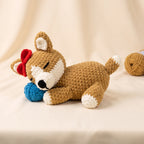 A crocheted plush dog toy in tan, white, and red, lying on a cream fabric background and holding a blue ball. A second ball of yarn sits blurred in the background.