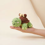 A small crocheted turtle plush toy being held in a person's hand against a soft beige studio background.