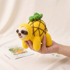 A hand holds a crocheted pineapple sloth toy against a soft cream fabric backdrop. A small printed card and crochet tools are visible nearby.