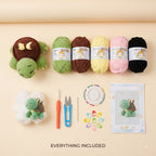 A flat lay of a crocheted turtle amigurumi kit showing a finished green and brown turtle, five balls of yarn in green, brown, pale yellow, pink, and black, crochet hooks, scissors, color swatches, stitch markers, and a printed pattern card on a beige background with 'EVERYTHING INCLUDED' text.