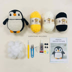 Crochet penguin kit laid out on a light beige background, including a crocheted penguin, balls of yarn in black, white, and yellow, stuffing, crochet hook, scissors, pins, glue, buttons, and a penguin crochet pattern booklet.