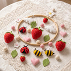 A handmade felt baby mobile or garland displayed on a round wooden hoop over a lace doily. The decoration includes red strawberries, pink hearts, white clouds and flowers, green leaves, ladybugs, bees, and small pom-poms.