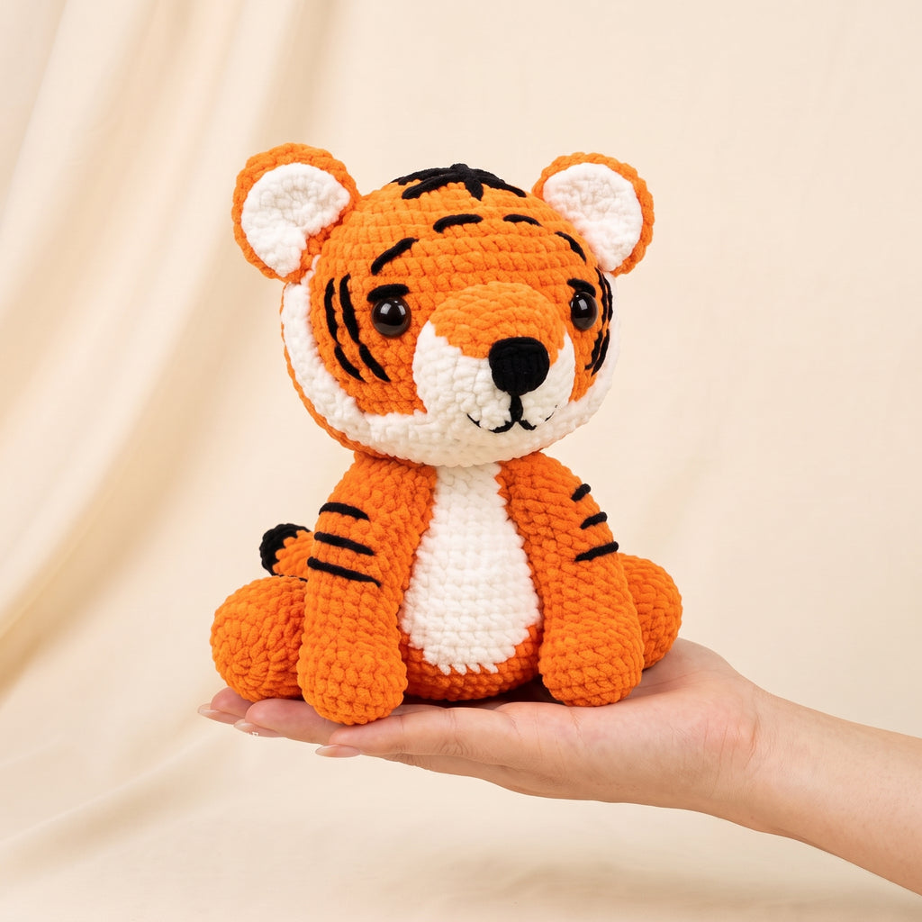A crocheted tiger plush toy with orange, white, and black yarn, being held in a person's hand against a soft beige fabric backdrop.