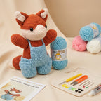 Crocheted fox plush toy wearing light blue overalls, displayed with a crochet kit pouch, yarn balls in blue, pink, and white, and a printed instruction card on a beige fabric background.