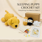 A crocheted puppy sleeping pillow kit displayed on a beige fabric background with its contents arranged neatly. The set includes a yellow crocheted puppy, a brown cardboard box, two balls of yarn (one tan, one black), white stuffing, a small beige drawstring pouch with crochet tools, colorful stitch markers, and a printed instruction booklet.