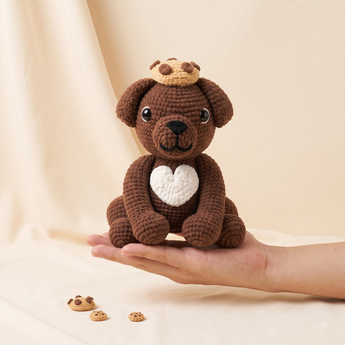 A brown crocheted plush puppy toy with a white heart on its chest and a small cookie crown sits on a person's hand against a warm beige fabric backdrop.