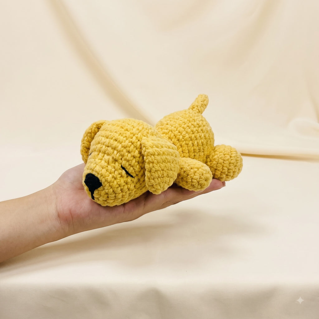 A small crocheted yellow puppy toy with a black nose and closed eyes, resting on a person's hand against a soft beige fabric backdrop.