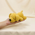 A small crocheted yellow puppy toy with a black nose and closed eyes, resting on a person's hand against a soft beige fabric backdrop.