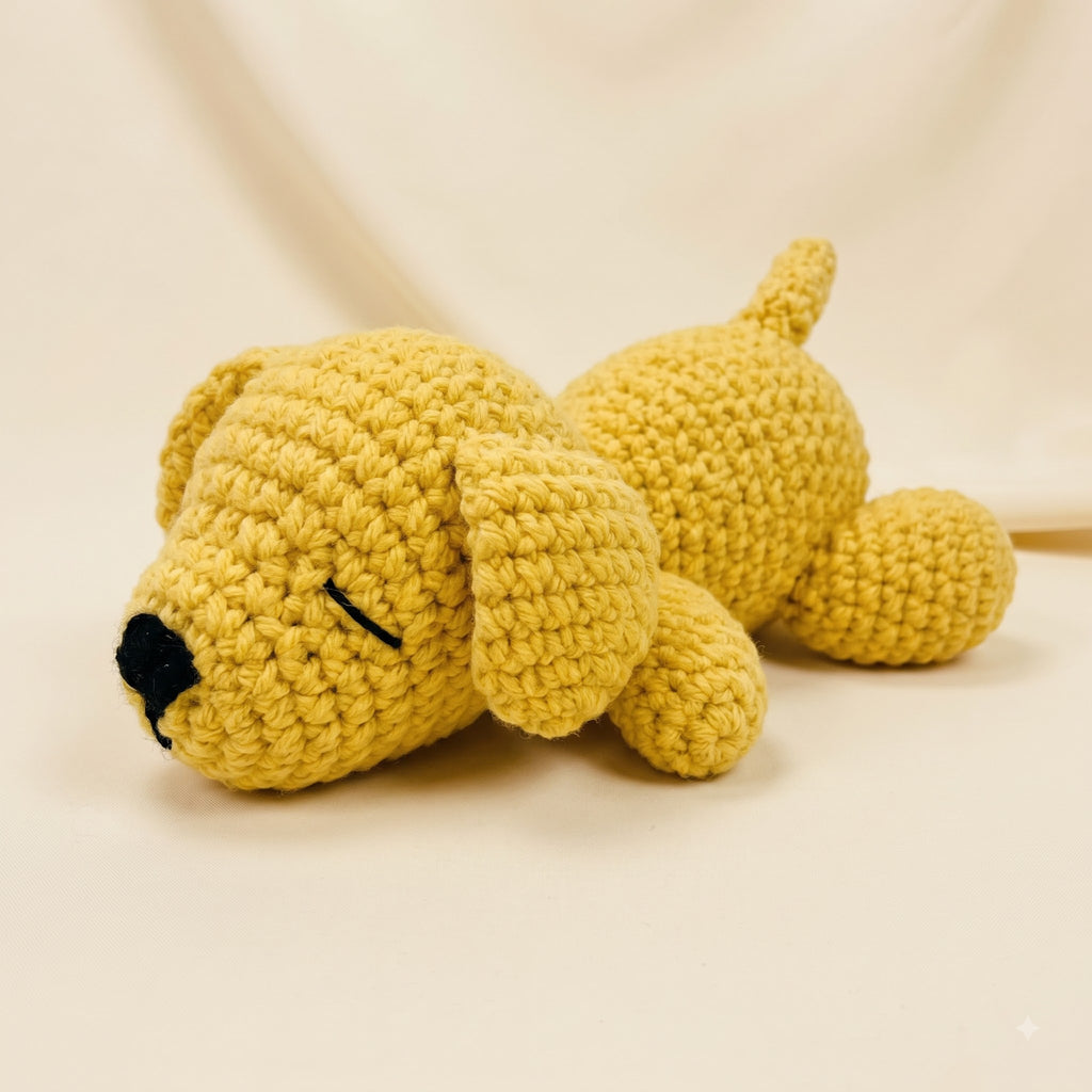 A yellow crocheted plush puppy toy with a black embroidered nose and closed eyes, lying down on a light beige background.