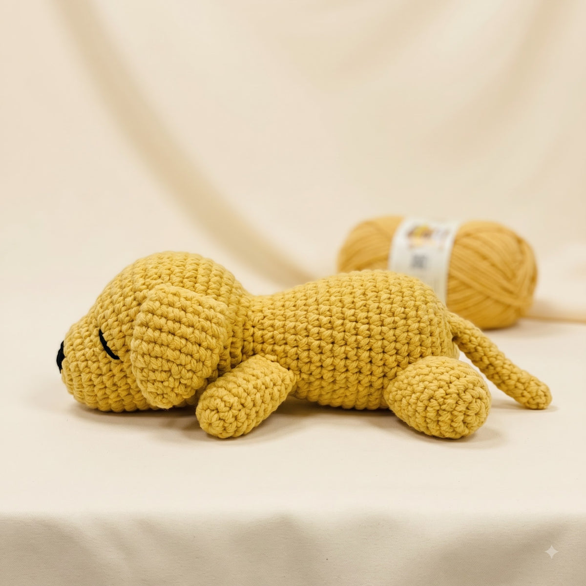 A yellow crocheted plush puppy toy lying on a cream fabric surface, with a matching yellow yarn ball blurred in the background.