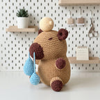A crocheted plush toy bear with a tan body, dark brown ears and feet, and light blue teardrop-shaped accessories, posed on a white surface against a white pegboard wall with shelves and small decor items.