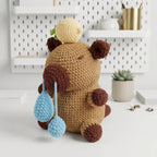 A crocheted plush toy resembling a bear or duckling in tan and brown, with a cream shell on its head, hanging light blue teardrop shapes, and a small green leaf detail, displayed on a white tabletop with shelves and a small plant in the background.
