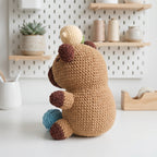A crocheted plush bear toy sits on a white tabletop in a bright, styled room. The bear is tan with dark brown ears, paws, and feet, holding a small cream-colored mushroom and a blue ball.