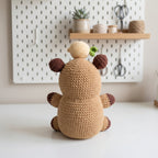A crocheted plush toy shaped like a small bear or animal character, shown from the back on a white tabletop. The main body is warm beige/tan, with darker brown ears, paw pads, and tail, plus a small green leaf detail on top.