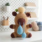 A crocheted plush toy bear sits on a white tabletop in a styled room setting. The bear is tan with dark brown ears, paws, and feet, a brown snout, and a small green leaf on its head.