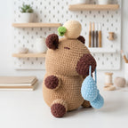 A crocheted plush toy bear in tan, brown, cream, and light blue, sitting on a white surface with a softly blurred craft room background.
