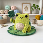 A crocheted green frog toy holding a dark green shamrock sits on a round green leaf-shaped mat on a white tabletop. The background shows shelves with yarn balls, a small plant, and framed art.
