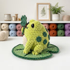 A crocheted frog toy sitting on a green leaf-shaped mat, with yarn balls and framed art in the background.
