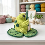 A crocheted frog toy sitting on a leaf-shaped cushion on a white tabletop, with yarn balls, a small plant, and shelves in the background.