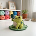 A crocheted frog toy sitting on a round green mat, holding a small four-leaf clover. The background shows a white shelf with colorful yarn balls and a small potted plant.