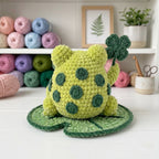 A crocheted frog plush toy in light green with darker green spots, sitting on a green lily pad and holding a four-leaf clover, set on a white tabletop with shelves of colorful yarn and home decor in the background.