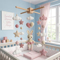 A pastel pink and cream crocheted baby mobile with a bunny plush, stars, balls, and clouds, hanging over a white crib in a light blue nursery room.