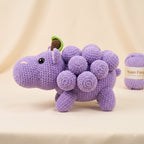 A crocheted purple dinosaur toy with a leaf on its head, displayed on a beige fabric backdrop next to a ball of matching purple yarn. The yarn label reads 'Nuan Fong'.