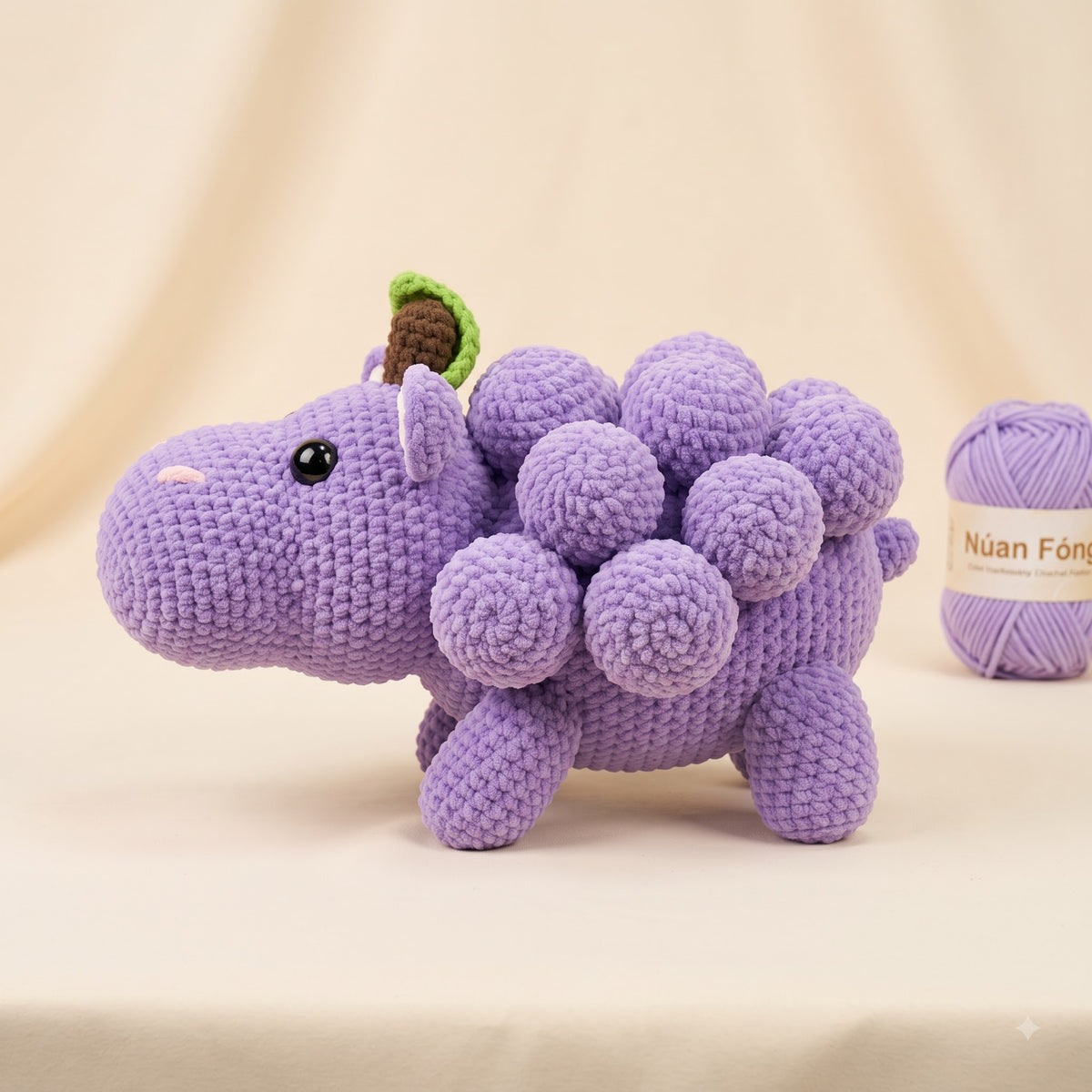 A crocheted purple dinosaur toy with a leaf on its head, displayed on a beige fabric backdrop next to a ball of matching purple yarn. The yarn label reads 'Nuan Fong'.