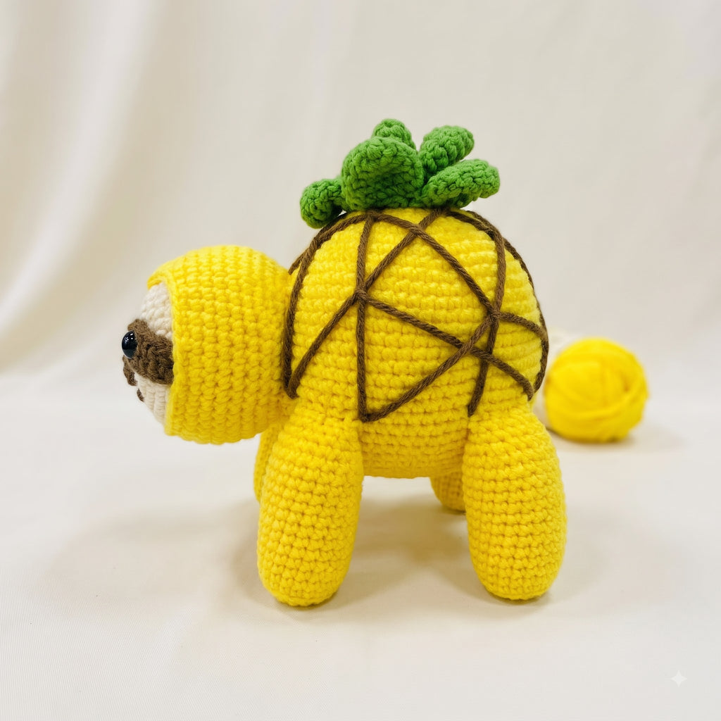 A yellow crocheted plush toy shaped like a pineapple, standing on a white fabric background with a small yellow ball behind it.