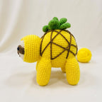 A yellow crocheted plush toy shaped like a pineapple, standing on a white fabric background with a small yellow ball behind it.