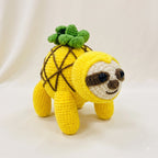 A crocheted plush toy shaped like a sloth wearing a pineapple costume, featuring a bright yellow body, green leafy top, and brown facial details, photographed against a soft cream fabric backdrop.