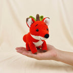 A small crocheted fox toy with a brown antler-like crown and green leaves is held in a person's hand against a soft cream fabric backdrop.