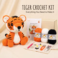 Tiger crochet kit displayed on a beige fabric background, featuring a crocheted tiger plush, a pattern booklet, a cardboard box, orange and white yarn balls, black yarn, crochet hooks, thread, scissors, and other small crafting tools. Text at the top reads 'TIGER CROCHET KIT' and 'Everything You Need to Make It'.