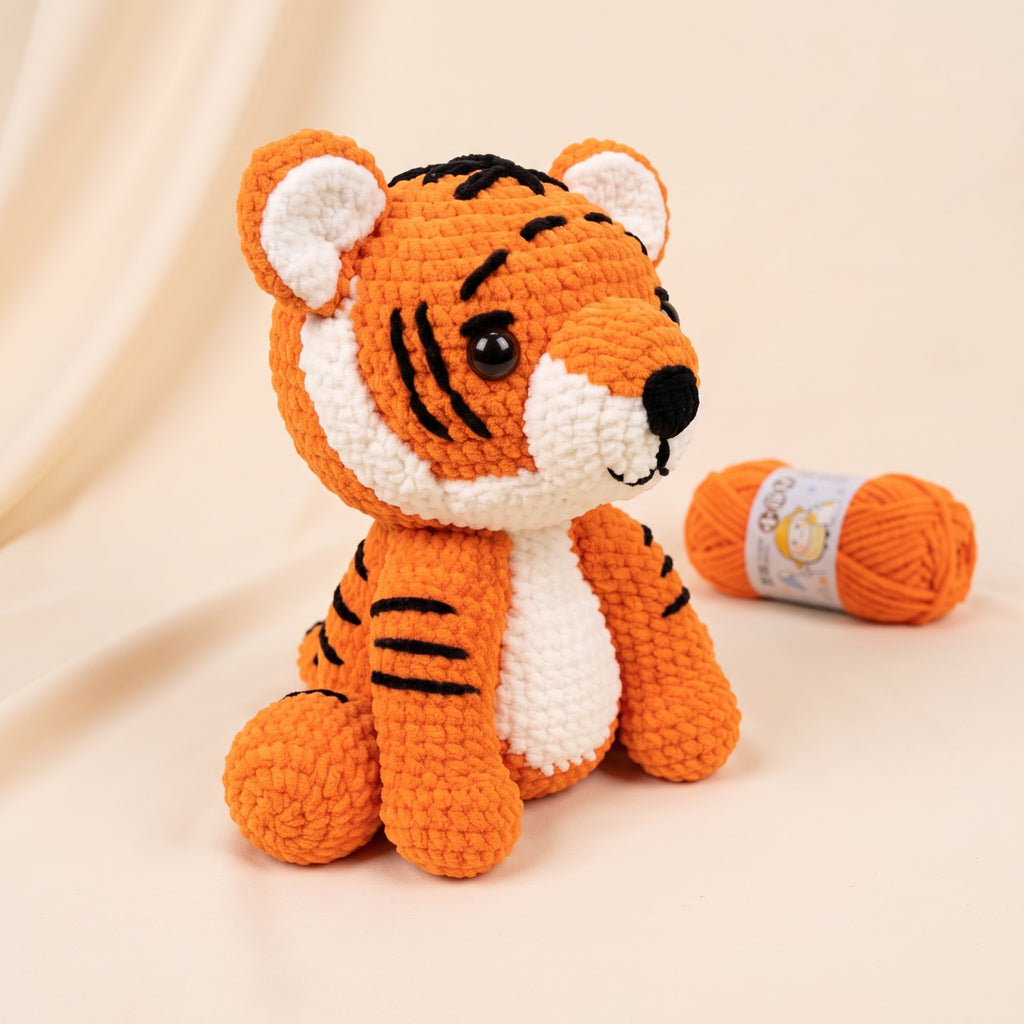 A crocheted tiger plush toy in orange and white with black stripes, posed next to a matching orange yarn ball on a warm beige background.
